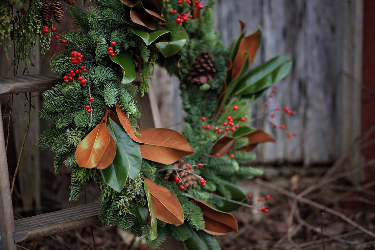 A handmade green wreath featuring magnolia leaves, red berries and pinecones in front of a barn backdrop.