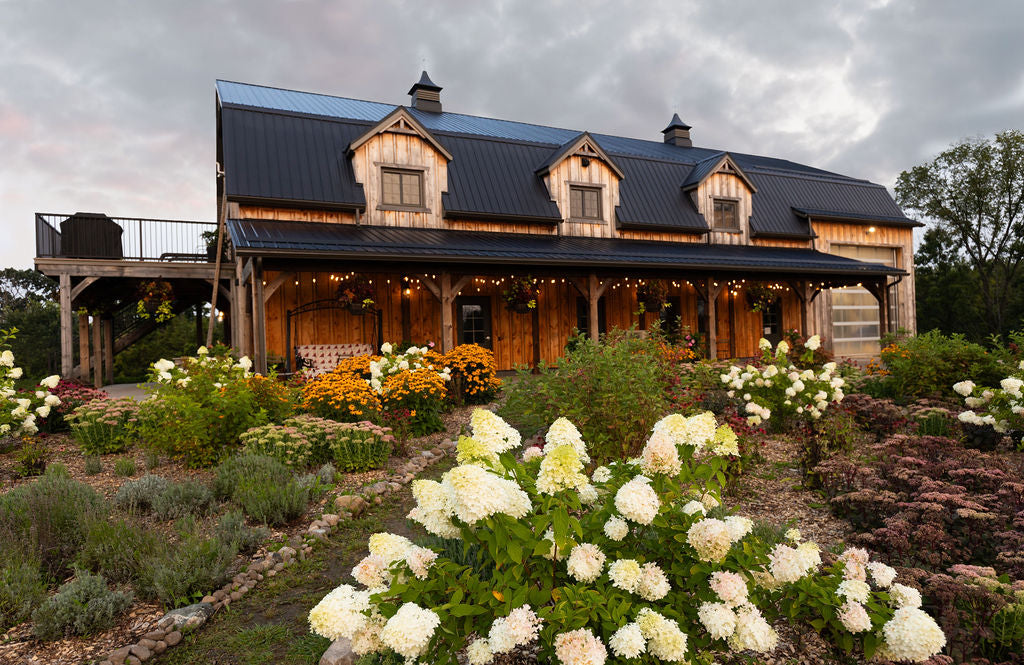 A wooden barn with a black wooden roof behind a large, perennial garden.