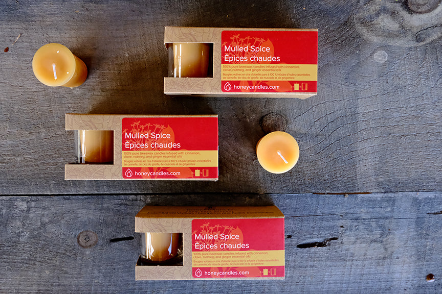 Three votive candles with red labels 'Mulled Spice Beeswax Votive Candles' in open cardboard packaging, displayed on a wooden surface.