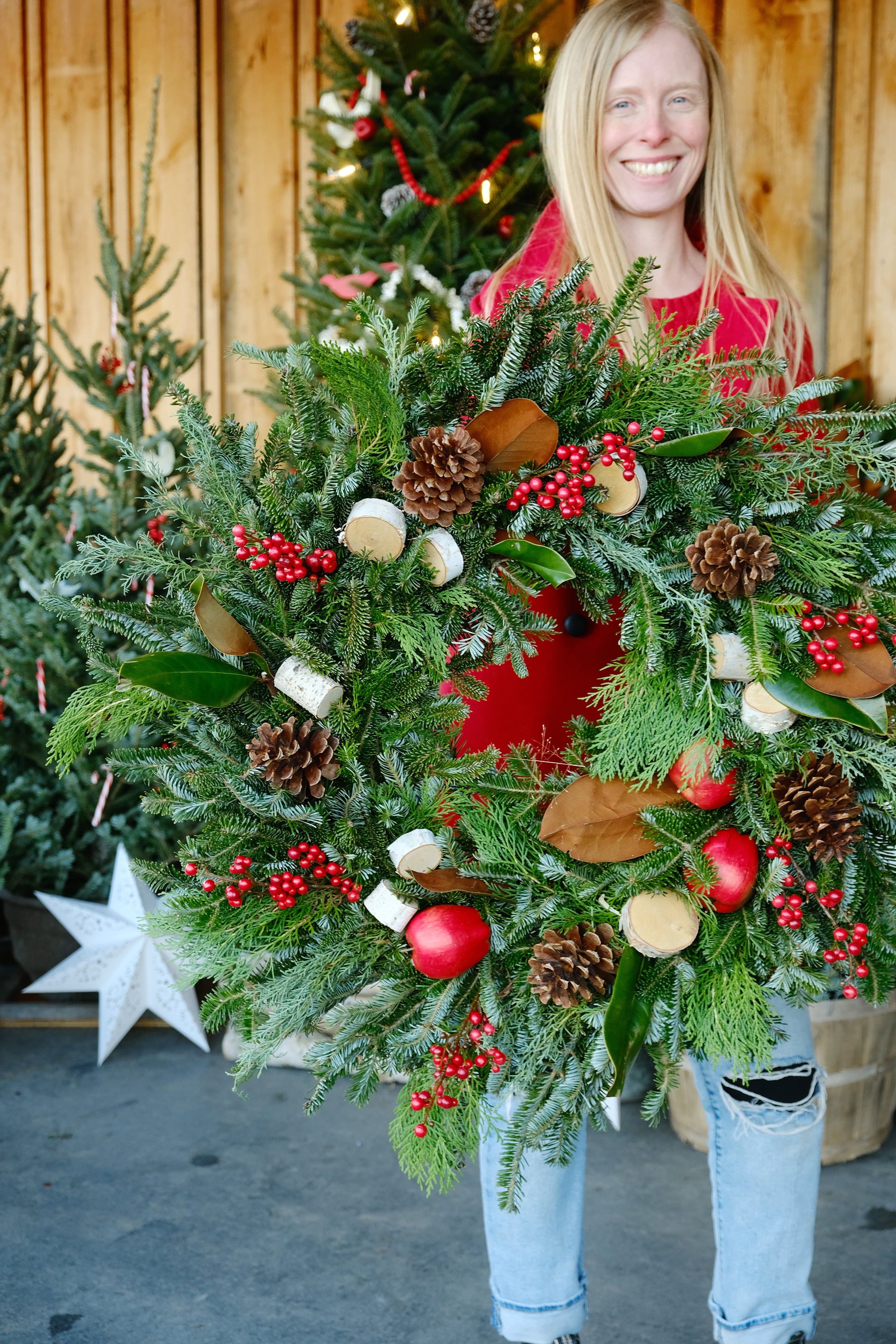 A person standing outdoors wearing a red coat and blue jeans, holding a handmade wreath with a combination of green evergreens, red berries, and other Christmas decorations.