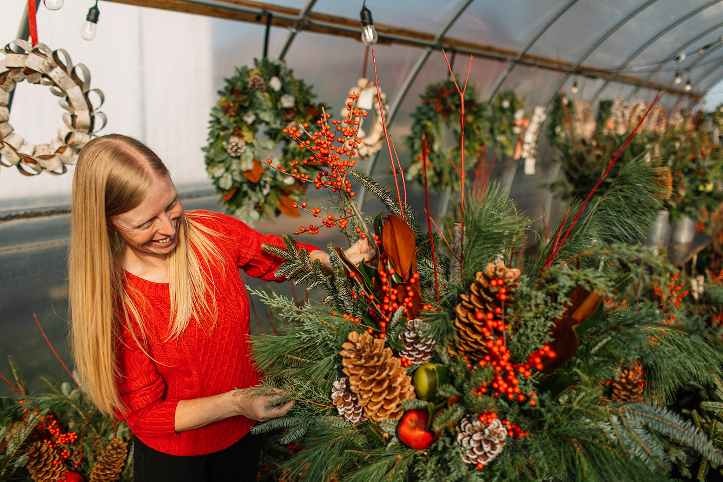 Someone arrange greens in a planter with berries, dogwood branches, red berries, pinecones with the backdrop of a greenhouse filled with other planters.