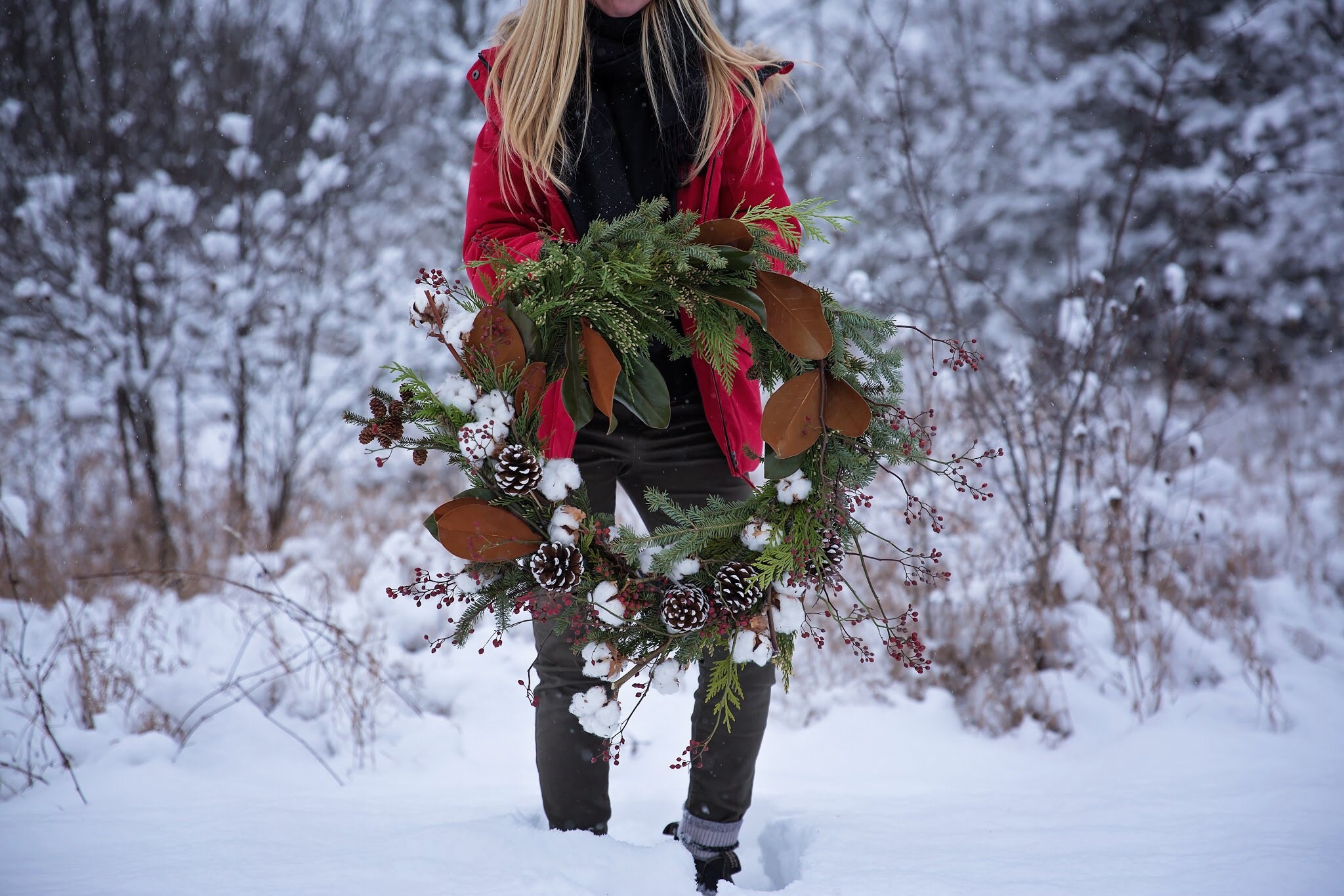 Holding a handmade wreath featuring winter greens, pinecones, magnolia leaves, red berries and cotton stems in the snow.