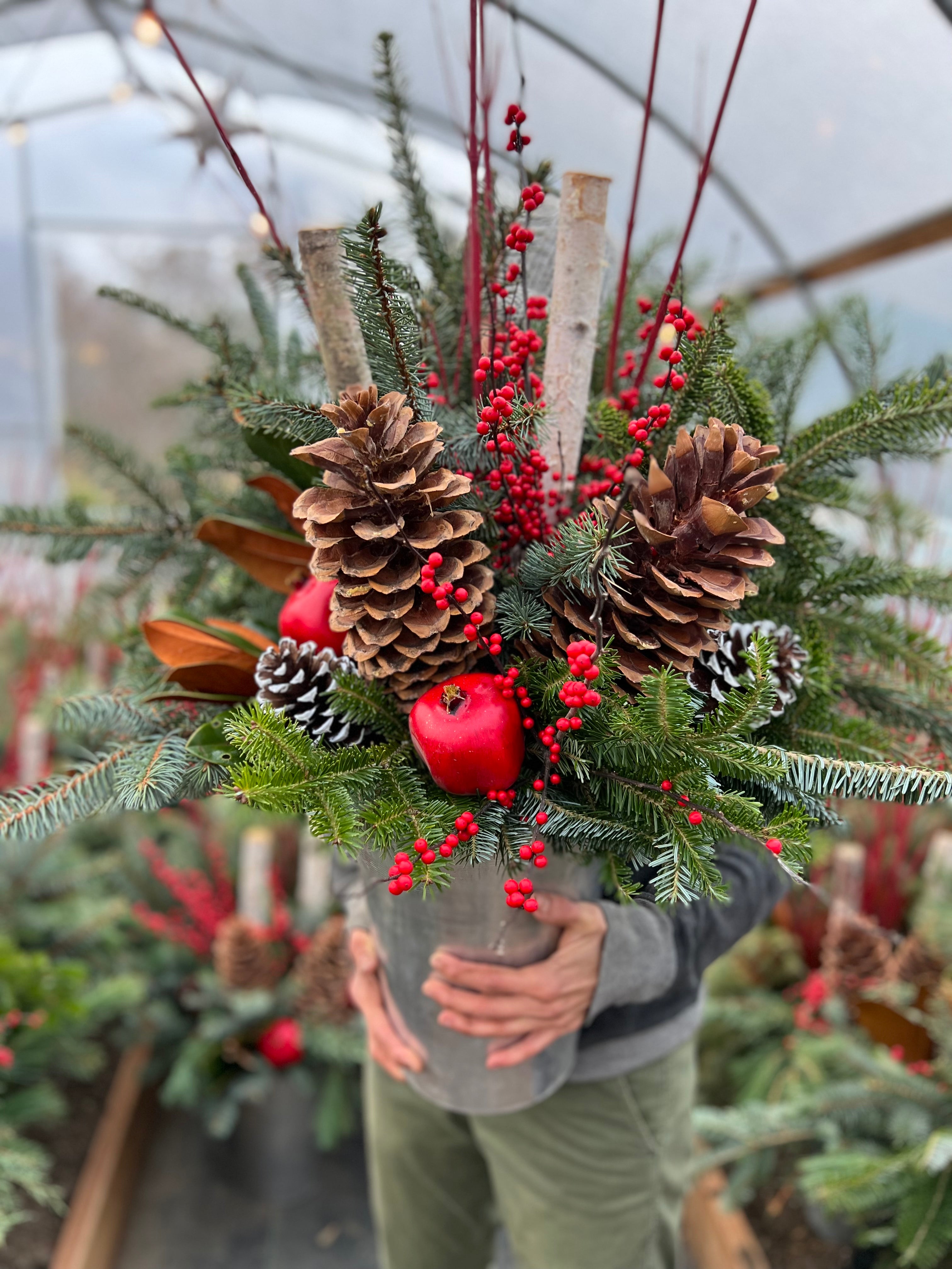 Someone is holding a metal sap bucket filled with various winter greens, birch poles, large pinecones, magnolia leaves, red berries and fruit.