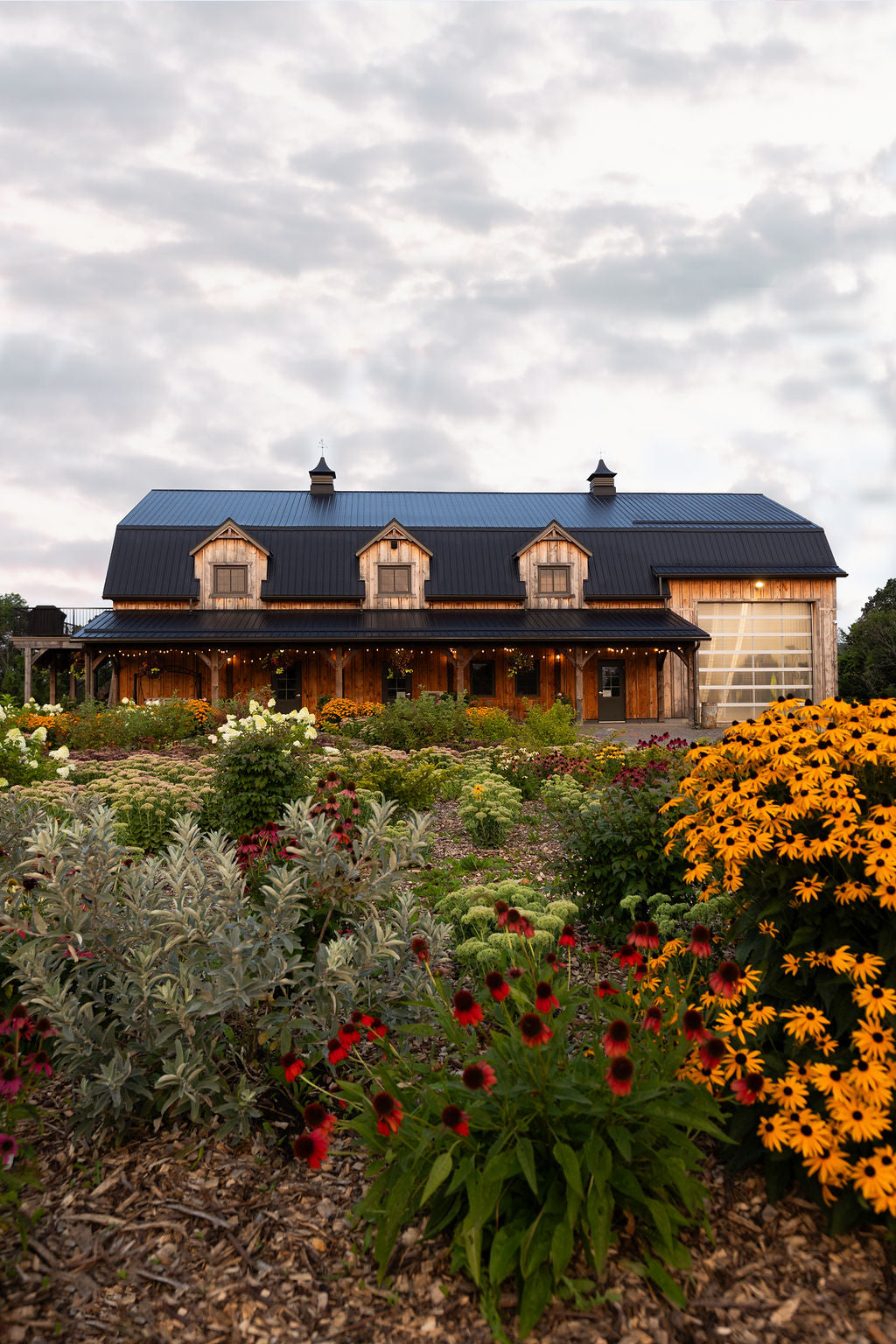 A wooden barn with a black roof and large windows, surrounded by a colourful flower field with predominantly yellow flowers in the foreground.