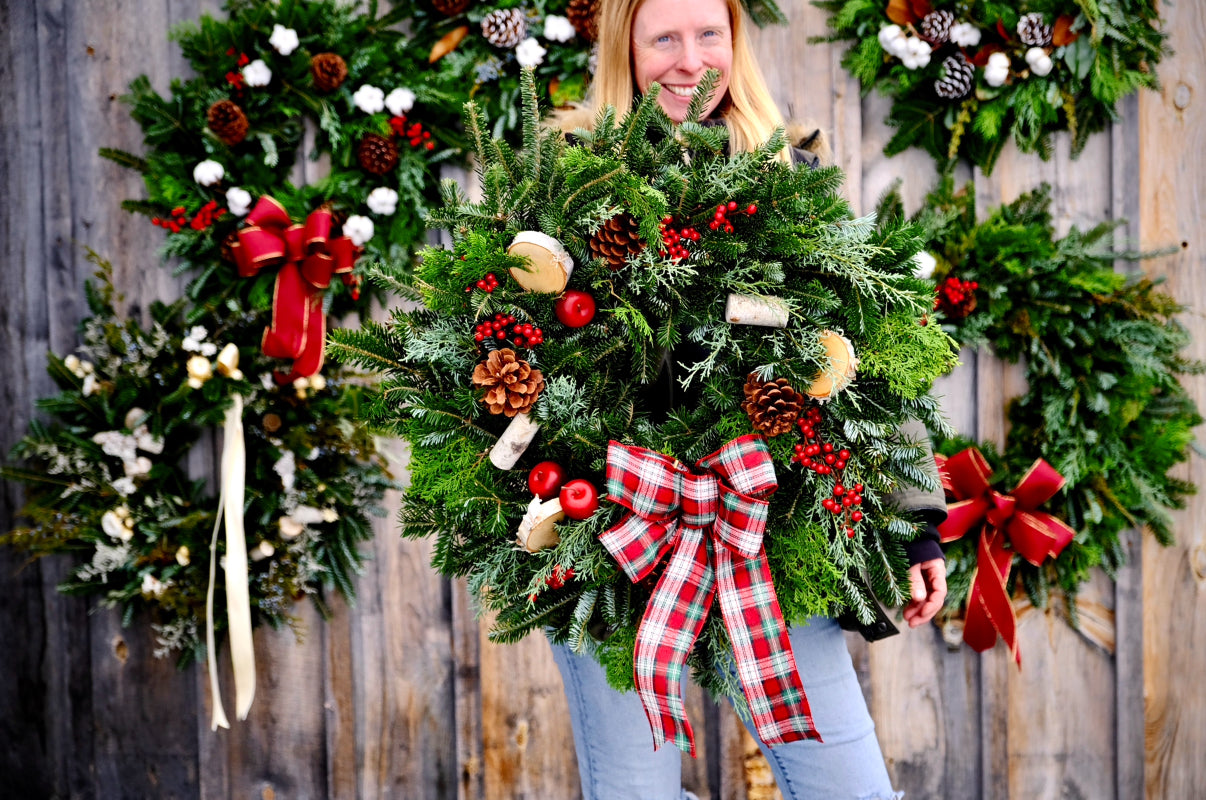 Person holding a large decorative wreath with other wreaths in the background on a wooden wall.