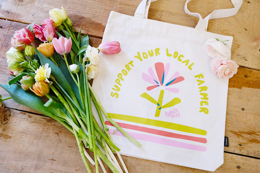 Off-white canvas bag with a flower and support your local farmer on it. On a wooden background with a bouquet of tulips beside it.