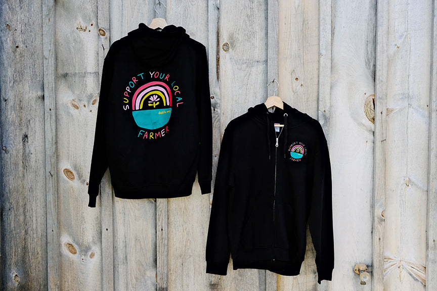 Two black hoodies with full zip, one displayed in front of the other, against a wooden backdrop. Both feature graphic prints on the front and back.
