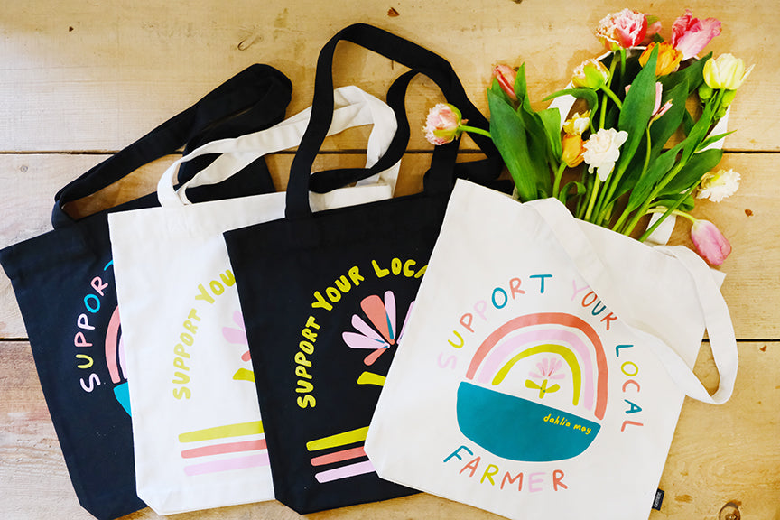 Canvas tote bags with the phrases 'Support Your Local Farmer' and 'Shop Local' printed on them, displayed alongside a bouquet of flowers.