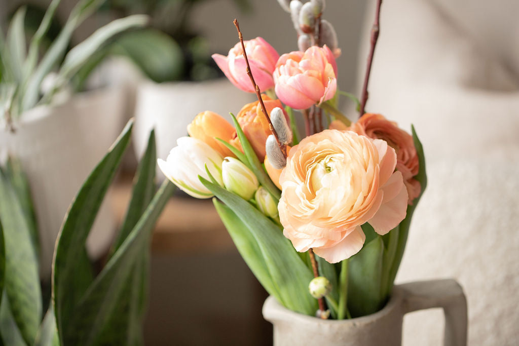 Close-up of a bouquet in a ceramic jug featuring ranunculus, tulips and pussy willow in peach and light pink colours.