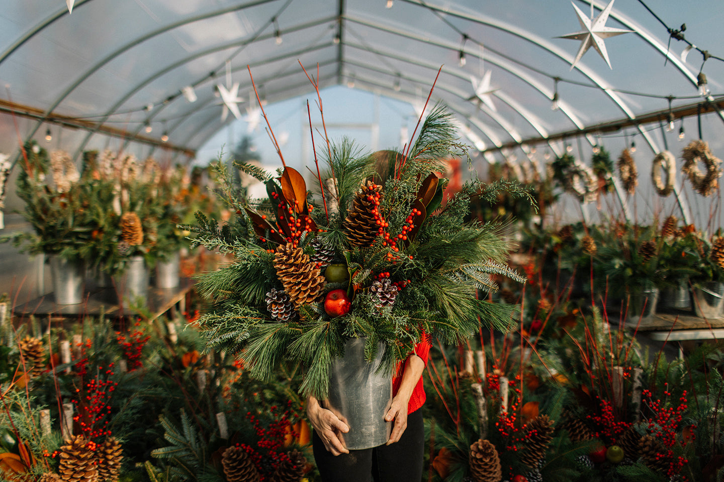 A person standing in a greenhouse holding a winter planter in a metal sap bucket. The planter features a variety of greens, pine cones, red dogwood, red berries and fruit.