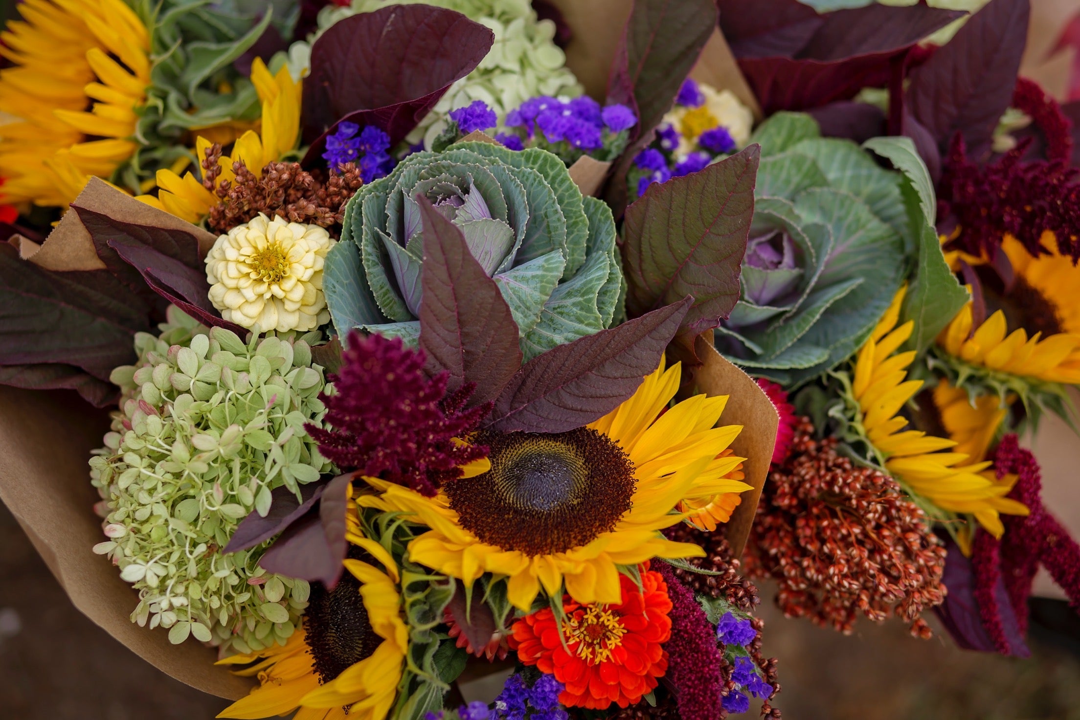A hand-tied bouquet with a variety of flowers including sunflowers and other seasonal blooms, wrapped in kraft paper.