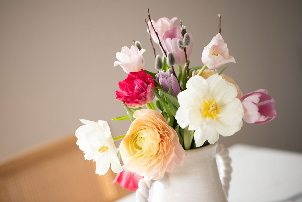 Close-up of a bouquet in a ceramic jus featuring peach ranunculus, white, fuschia and light purple tulips.