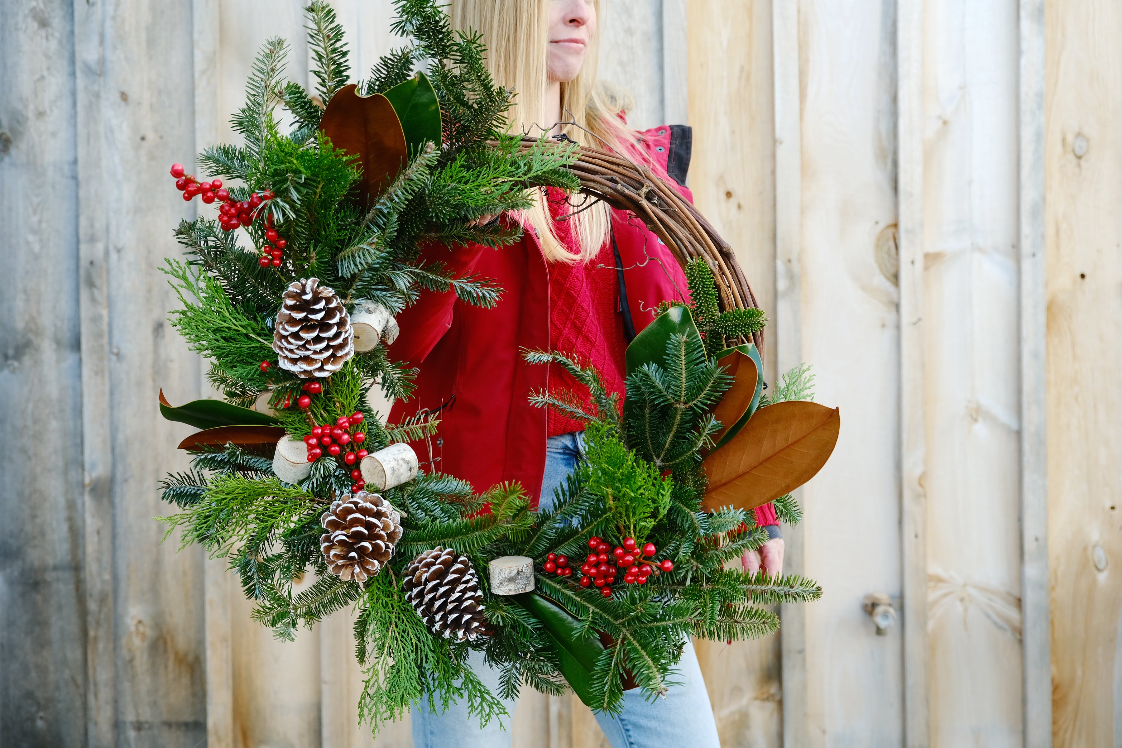 A handmade asymmetrical grapevine wreath with a variety of fresh evergreens, berries, and other natural elements, held in front of a wooden fence.