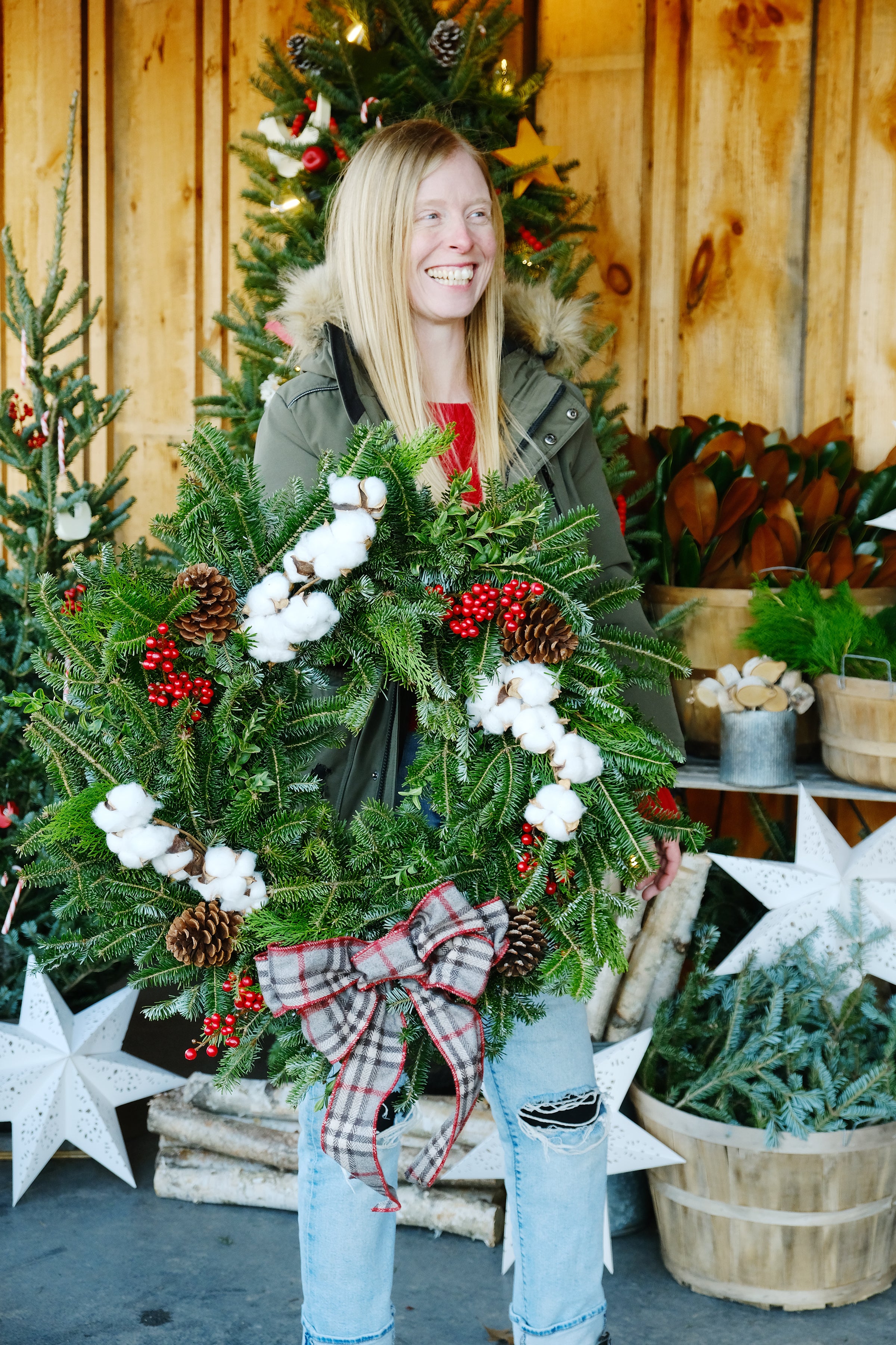 A person standing in a barn-like setting with a Christmas wreath made of evergreens, berries, and a plaid bow in the center, with decorative stars and a Christmas tree in the background.