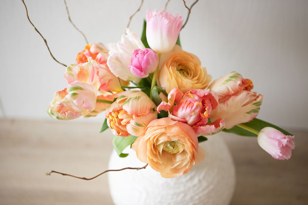 Close-up of bouquet in a ceramic vase featuring peach ranunculus, light pink and white tulips and curly willow. 